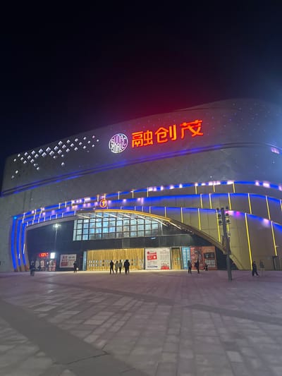 A tall building with a curved facade is illuminated at night. The building features blue and yellow lights, and a large red neon sign in Chinese characters runs vertically along its side. A number "1" is visible near the entrance. People are standing and walking on the tiled walkway in front of the building, which is also brightly lit. The sky is dark.