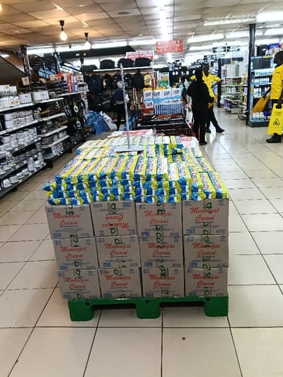 The image captures an interior view of what appears to be a supermarket or large retail store aisle. The central focus is a large product display set up on a green plastic pallet on the tiled floor.
In the immediate foreground and center, numerous cardboard boxes are stacked on a green, short plastic pallet. These boxes form the base of the display. Many of the boxes prominently feature the brand name "Menengai Cream" printed in a reddish-brown font. Below the brand name, some boxes read "QUALITY STANDARD ISO" and "20 KG" (or similar weight indications like "20 X KG"). A heart symbol is also visible on several boxes. Some boxes are oriented upside down or sideways, showing the text inverted or on different sides.
Stacked on top of these cardboard boxes are multiple layers of individually packaged items. These packages are primarily yellow and blue, arranged horizontally in rows, creating a large, flat surface. The wrappers appear to contain a product consistent with "Menengai Cream," likely a type of cooking fat or cream.
To the left of the central display, there are extensive metal shelving units stocked with various items. These shelves hold numerous silver-colored containers, pots, or kitchenware, suggesting a household goods or hardware section. Further back on the left, more shelves and products are visible, along with what looks like black bags or backpacks hanging from a display.
In the mid-ground and background to the right of the central display, an aisle extends further into the store. Several people are visible in this area. At least two individuals are wearing bright yellow shirts and dark trousers, suggesting they are store staff or employees. One of the staff members in yellow is holding a yellow object, possibly part of a "wet floor" caution sign. Another yellow caution sign, featuring a black silhouette of a person slipping, is visible near the right edge of the frame. A third person, dressed in black, is also present, possibly interacting with the staff or browsing. Shelves stocked with various products line the right side of this aisle.
Overhead, the ceiling is fitted with fluorescent lighting fixtures, illuminating the store. Several hanging signs are visible in the upper mid-ground. One prominent red sign reads "QUICK MART" with additional smaller text below it. Another sign further back appears to indicate departments, with some visible text like "FURNITURE & ELECTRONICS" and "DETERGENTS" (partially visible). The floor is made of light-colored tiles.
The overall scene depicts a busy retail environment during what appears to be regular operating hours, with products displayed for sale and staff present.
