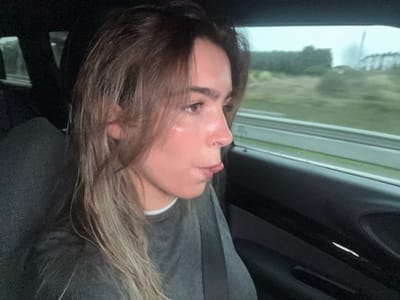 Inside a car, a young woman with shoulder-length, light-brown hair, partially wet, sits buckled. She wears a gray sweater. Her face is turned to the right, sticking her tongue out slightly. The background shows blurred green foliage and a cloudy sky visible through the car window, suggesting movement. The interior of the car, with dark upholstery, partially frames the scene.