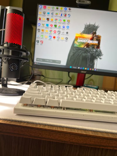 This static image, captured in Donchevo, Bulgaria, presents a close-up view of a computer workstation. In the foreground, a white mechanical keyboard is visible, with several keycaps removed from the left side, exposing brown mechanical switches and accumulated debris. The keyboard rests on a white mousepad, which in turn sits on a wooden desk surface. To the left of the keyboard, a black HyperX QuadCast S microphone stands, illuminated with a distinct red honeycomb pattern. In the background, an Acer computer monitor displays a desktop environment. The screen shows numerous application icons arranged on the left side, and a desktop wallpaper featuring a dark, cloaked, crowned, and veiled figure. An image of "Ghost Recon" game art is overlaid on the wallpaper. A Logitech webcam is mounted centrally on the top edge of the monitor. The background behind the monitor consists of a blurred greenish wall with indistinct objects. No individuals are present, and no actions are in progress.