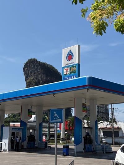 The image captures a PTT gas station in Phang-nga, Thailand. The station has a blue and white canopy with red accents. The PTT station's sign features the company logo and the logos of 7-Eleven and Amazon Cafe, indicating the presence of these businesses within the station. Gas pumps are visible under the canopy, with signs indicating fuel types (Diesel, E20, Gasoline 91, and Gasoline 95) and possibly prices. A sign with a handicap symbol is also present. The cashier area can be seen in the bottom of the image. In the background, a limestone karst mountain stands out against a clear blue sky.