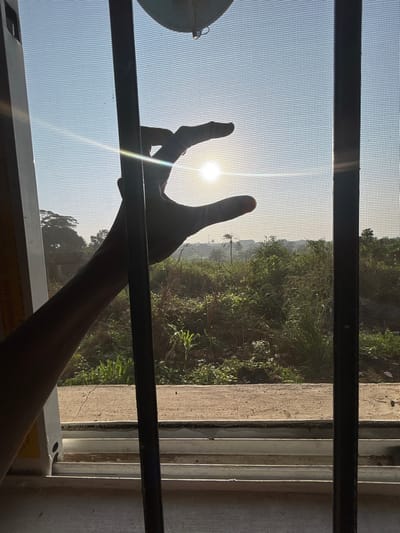 This image, captured in Debasco, Nigeria, depicts an outdoor scene viewed through a window from an interior vantage point. The window features a fine mesh screen and two dark vertical security grilles. A human left hand is silhouetted in the upper-left, with its index finger and thumb curled to partially frame a brilliant, low-lying sun, which emits strong lens flare across the frame. The background consists of a clear, light blue sky above a sprawling, dense landscape of green vegetation and trees. A slender palm-like tree is visible in the distant mid-right. The window frame and a weathered sill are present in the foreground at the bottom. A light-colored, curved object is partially visible at the top edge of the frame.