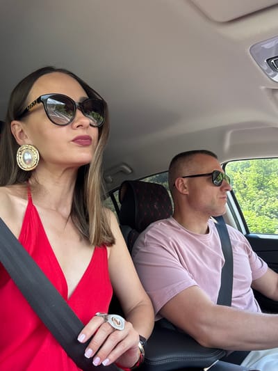 In the city of Chvizhepse, Russia, a man and a woman are seated in the front seats of a car. The woman, on the left, is wearing a red halter top and large, decorative earrings. She has on oversized sunglasses with gold accents and is wearing lipstick. A large, modern ring adorns her right hand, and her fingernails are painted a pale pink. A black seatbelt crosses her chest.

The man, on the right, is wearing a pink t-shirt and sunglasses. A black seatbelt is visible across his chest as well. His hair is closely cropped. Both individuals are looking forward, suggesting they are driving or observing the road ahead. The interior of the car is visible, with a patterned headrest of the seat behind the man. Through the car window on the right, lush green trees and foliage are visible, indicating an outdoor, possibly rural or forested, setting. The lighting suggests it is daytime.