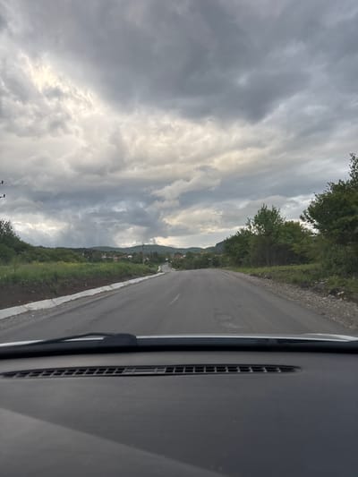 The image depicts a view from inside a vehicle, looking out onto an asphalt road in Baylovo, Bulgaria. The road stretches into the distance, flanked by vegetation on both sides, including trees and grassy areas. In the background, a village or town is visible near a hillside. The sky is overcast with gray and white clouds. The vehicle's interior, including a vent and windshield wiper, is visible on the right side of the image.
