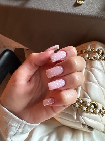A hand with long, square-shaped nails is prominently displayed, with a light pink base color and white polka dots. The hand is holding a beige quilted purse with gold chain straps. The background includes portions of a beige garment with a visible button, and an indistinct dark object. The scene is an indoor setting. The location is not discernible from the image.