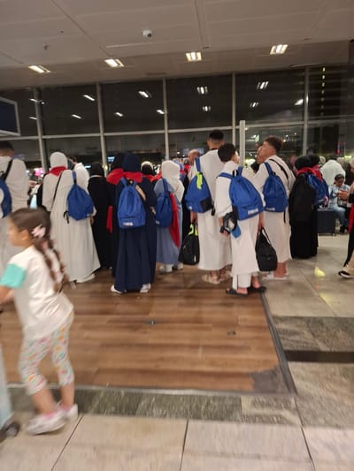 In Pendik, Turkey, a group of people, some wearing white ihram clothing and red scarves, are gathered at what appears to be an airport or transportation hub. Several individuals, mostly young men and women, are wearing blue backpacks. A young girl in a white shirt and patterned leggings is visible in the foreground, walking past. The environment features large windows, fluorescent lighting, and a transition from a wooden floor to tiled flooring. The overall scene suggests a moment of waiting or transit.