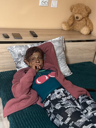 An elderly woman lies on a bed in what appears to be an apartment setting. She is positioned on a pillow with a gray and white pattern, covered by a green textured blanket. She wears a dark teal shirt with a cherry design, a pink cardigan, and a patterned black and white robe. Her hand is resting on her chin. A teddy bear is situated on the wall above a wooden cabinet, where a smartphone and other devices are placed. The wall is light beige with two electrical outlets. The location is likely a residential interior in Vinitsa, Bulgaria.