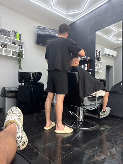 This media file captures an indoor scene within a modern barbershop, likely located in Simitli, Bulgaria. The image is taken from a low-angle perspective, showing a client reclining in a black barber chair with their head back, covered by a black cape. A barber, dressed in a black t-shirt and black shorts, stands behind the client, seemingly preparing for or in the process of washing the client's hair, though the sink is only partially visible.
The barbershop features a sleek, contemporary design with a black textured wall on one side and clean white walls on the other. The floor is made of glossy black marble or tile, on which scattered hair clippings are visible, indicating recent haircuts. Geometric fluorescent strip lights illuminate the ceiling and parts of the wall, reflecting in a large mirror that shows an air conditioner and some shelving units.
Notable details include the client's mixed footwear: one foot wears a multi-colored athletic sneaker, while the other is in a light-colored slip-on sandal. In the immediate foreground, the photographer's foot, clad in a distinctive white sneaker with black scribbled patterns, is visible. White shelving units adorned with various hair care products, bottles, and a small potted plant line the white wall. There is also a television mounted on the white wall. The overall ambiance suggests a professional and clean but busy environment. No distinct facial expressions or visible text are clearly legible.