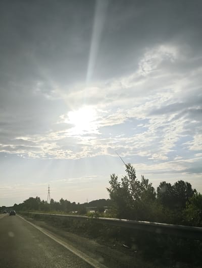 The image depicts a view from a moving vehicle on a highway. The foreground shows the asphalt road with a white lane marker, bordered by a metal guardrail and vegetation. In the background, trees and bushes line the side of the road, with power lines and electrical towers visible in the distance. Two cars are driving on the highway ahead.
The sky is dramatic, filled with a mix of dark and light clouds. A strong beam of sunlight cuts through the clouds from the upper left, illuminating the scene with a bright glow. The interplay of light and shadow suggests either an approaching storm or the clearing after one. The time of day appears to be late afternoon or early evening, given the quality of the light. The overall mood is somewhat somber yet awe-inspiring due to the celestial display. The location is identified as Teverola, Italy, implying the scene is set in the Italian countryside or outskirts. There is no visible text in the image.