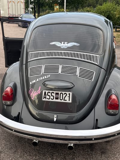 A dark gray Volkswagen Beetle is parked on asphalt in Skrea, Sweden. The car's rear is facing the camera, and the driver's side door is open. The car has several stickers on its rear: a winged emblem with the letters "LS" at the top of the rear window, the text "VW 1302" below it, and a pink cursive sticker that reads "Bad" to the left of the license plate. The license plate is visible and reads "ASS 021". A trailer truck and a blue car are parked behind the Beetle. The environment appears to be an outdoor parking area with trees and vegetation in the background.