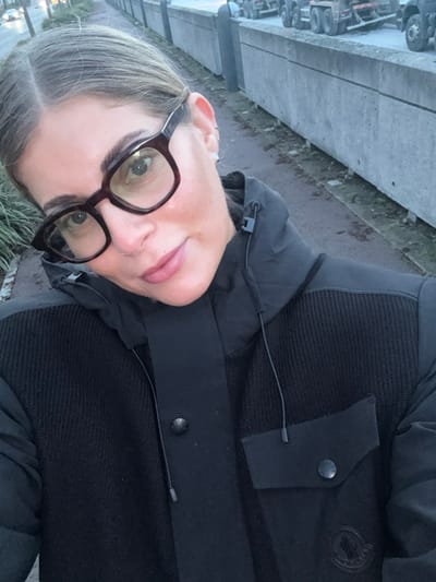 A woman with light hair and dark-framed glasses is the focus. She is wearing a dark jacket with buttons and a small flap. The background features a concrete structure with what appears to be parked vehicles. The overall setting appears to be an outdoor urban area.