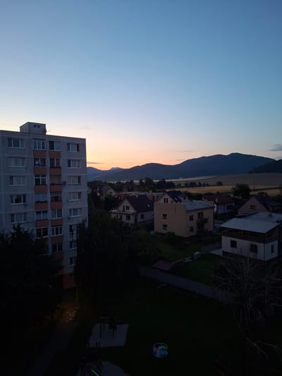 The image depicts a twilight scene in Ilava, Slovakia. A tall, grey apartment building dominates the left foreground, its windows illuminated with soft light. In the middle ground, several residential houses with red-tiled roofs are nestled among trees. Beyond the houses, rolling hills form a dark silhouette against the pale orange and blue gradient of the dusk sky. A thin line of trees and a field of vegetation can be seen in the distance. The lower part of the image shows a dark courtyard with a playground. The overall impression is one of a quiet residential area at the end of the day.