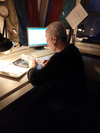 An older man with gray hair and a beard, wearing a dark long-sleeved shirt, is seated at a light-colored desk, viewed from behind and slightly to the right. He is actively engaged with a desktop computer, which features an older Samsung monitor displaying what appears to be a spreadsheet or data-rich document. A light-colored keyboard sits in front of the monitor. To the left of the man's hands, a black electronic calculator with the model number "SC2300" is positioned, along with several papers and what looks like a receipt.

The workspace is illuminated by artificial light, primarily from two desk lamps: a black adjustable lamp on the far left and a modern LED task light near the computer monitor. The background features a large flag with horizontal stripes, possibly the Russian flag, hanging on the wall. Above the black desk lamp, a circular analog gauge or clock is visible.

To the right of the man, a window is visible, reflecting internal lights. Attached to the wall or window frame is a clear plastic folder containing a printed document, possibly a checklist or set of instructions. The overall setting appears to be an indoor work area, potentially an office or control room, in Medvezhyegorsk, Russia, with the artificial lighting suggesting it might be evening or night.