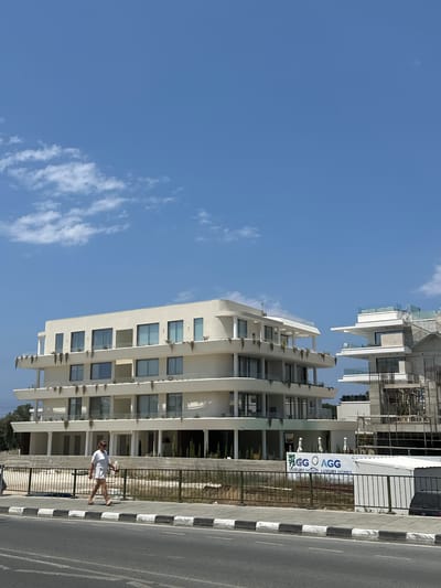 The image shows a street scene in Paphos Municipality, Cyprus, on a clear day with a blue sky and scattered white clouds. In the foreground, a person wearing a white outfit and sunglasses walks along a sidewalk next to a road. Behind the sidewalk, a decorative fence borders a construction site and a modern building complex. The building complex features several levels with balconies and large windows, painted in a light, cream color. To the right of this building, another structure is under construction, with exposed scaffolding and concrete elements. A banner displaying "AGG LUXURY HOMES" is visible in front of the main building. Trees and some greenery are present on the grounds of the complex. The road in the foreground has a striped white and black border.
