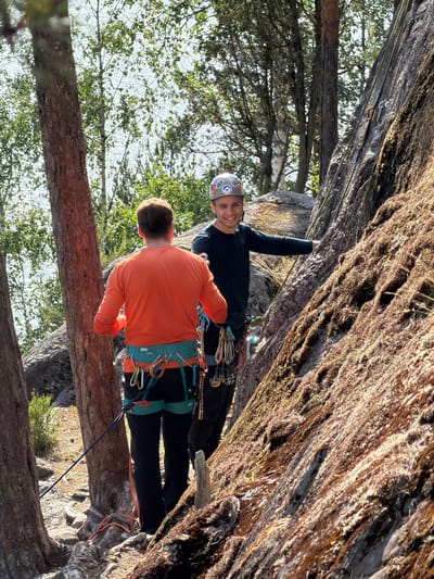 Two individuals are engaged in rock climbing near a body of water, likely a lake, in Ruch'i, Russia. The person in the foreground, wearing an orange long-sleeved shirt and black pants with a teal harness, is facing away from the camera, towards the rock face. They are equipped with climbing gear, including quickdraws attached to their harness. The second individual, in the middle ground, is wearing a dark long-sleeved shirt and a helmet with a patch, and is smiling at the camera, looking towards the rock. They are also wearing a harness and carrying climbing equipment. The rock face is covered in moss and vegetation, indicating a natural outdoor environment. Several trees are visible in the background, with the shimmering surface of water behind them. The ground is rocky and covered with pine needles and small bushes. The scene appears to be a sunny day, as indicated by the lighting and shadows.