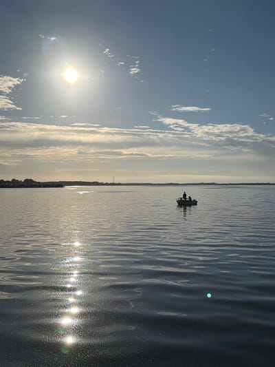 The media displays a wide shot of a body of water with a person or people in a small boat. The sun is visible in the sky, casting a bright reflection on the water's surface. The sky is partly cloudy, with the sun illuminating the clouds from behind. The water appears calm, with small ripples and a shimmering effect from the sunlight. The overall scene evokes a sense of peace and tranquility. The image is oriented vertically, with the sky on the left and the water on the right. A distant shoreline is visible, with trees and a wind turbine, suggesting a rural or coastal location. The time of day appears to be morning or late afternoon, given the angle of the sun. The weather seems clear and pleasant. No text is visible in the image. The location context provided is Ribnitz-Damgarten, Germany.