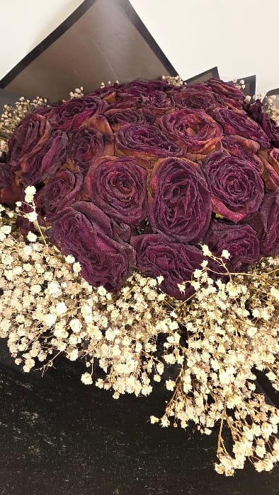 The image depicts a close-up, slightly overhead view of a preserved floral bouquet. The central arrangement consists of numerous dark red to burgundy roses, tightly clustered and exhibiting a dry, textured, or frosted appearance. These roses are accented and encircled by abundant clusters of small, white, dried flowers, consistent with *Gypsophila*. The bouquet is partially encased in dark, likely black, wrapping material visible at the bottom and along the sides. In the upper background, dark, angular elements suggest further packaging or a display box, set against a plain, light-colored surface. The image contains no identifiable landmarks, architectural features, or contextual details that could confirm its location in Heusden, Belgium, or any other specific geographic area.