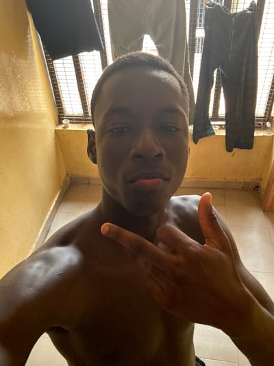 A dark-skinned male subject is depicted in a close-up, vertical-format image, shirtless and facing the camera directly. He has short, dark hair and a lean build, with his shoulders and upper chest visible. His expression is neutral to slightly pouting. His right hand is raised towards his chest, forming a gesture with the index finger and thumb extended, while the middle, ring, and pinky fingers are curled. The background consists of a light yellow-painted wall. Behind the subject's head and to his left, a window or opening secured by a metal grate is visible. Several items of clothing, specifically two pairs of dark trousers, are hanging on a line to dry against the grate. The floor, visible in the lower right corner, appears to be tiled. This scene is located in Ilishan, Nigeria.