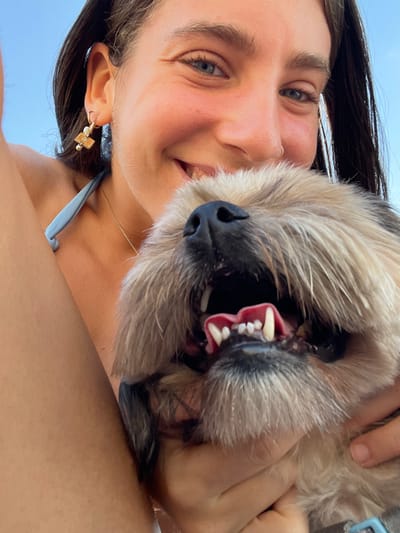 A close-up, low-angle image captures a young adult female holding a dog outdoors in Palmachim, Israel. The female, positioned in the upper portion of the frame, has dark hair, blue eyes, and a small nose ring on her left nostril. She is smiling, revealing her teeth, and wears a light blue bikini top with a gold-colored hoop earring featuring a rectangular charm on her left ear. Her tanned right arm is visible on the left. The dog, a small to medium-sized breed with shaggy light brown and dark brown fur, occupies the lower part of the frame. Its mouth is open wide, exposing pink gums and white canine teeth. Both subjects appear to be looking directly at the camera. The background is an undifferentiated clear, bright blue sky.