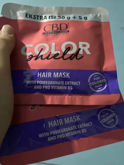 Two sachets of CBD Color Shield Hair Mask are visible. The top sachet has "EKSTRA ISI 30 g + 5 g" printed at the top, followed by "CBD CREATIVE BEAUTY DAZZLE PROFESSIONAL". Below this, "COLOR shield" is written in large font. A purple band across the sachet reads "HAIR MASK WITH POMEGRANATE EXTRACT AND PRO VITAMIN B5" and a circular emblem states "FOR COLORED HAIR". The bottom sachet is partially visible, showing a similar purple band with the text "HAIR MASK WITH POMEGRANATE EXTRACT AND PRO VITAMIN B5". The sachets are pink with purple and white text. A person's thumb and forefinger are holding the top sachet on the left side of the frame. The background is a light blue surface. The location is identified as Banyusri, Indonesia, however, this information is not visually derivable from the image itself.