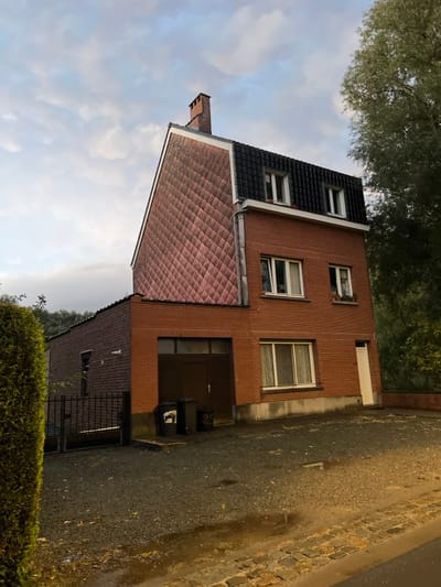 A three-story residential building, constructed primarily of red brick, is depicted under an overcast sky in Anderlecht, Belgium. The structure features a complex roofline: a steep, gabled section on the left is clad in diamond-patterned, weathered pinkish-red tiles and topped by a brick chimney. This section adjoins a mansard-style roof on the right, covered in dark, possibly black, tiles, featuring two white-framed dormer windows. A silver-colored vertical downspout or trim separates these two roof sections.
The main facade displays multiple windows: the ground floor has a large, horizontal, white-framed window with light-colored curtains. The first floor presents two white-framed windows; one double-pane window with curtains and a smaller window to its right, which has a window box containing green foliage beneath it. The ground floor also includes a dark brown garage door or access opening on the left, near which two dark refuse bins, one marked "25", are positioned. A narrow, white-framed entrance door is located on the far right of the ground floor.
To the left of the main building, a single-story brick extension with a flat, dark roof projects outwards. A dark metal gate is partially visible in front of this extension. The immediate foreground consists of an unpaved, gravel or crushed stone surface exhibiting multiple puddles, suggesting recent precipitation. This surface leads to a cobblestone curb in the very foreground. A dense, trimmed green hedge borders the left side of the scene, partially obscuring the extension. A large deciduous tree with light green foliage is visible on the far right, partially framing the upper portion of the house. No human figures or animals are present. The ambient light suggests either dawn or dusk.