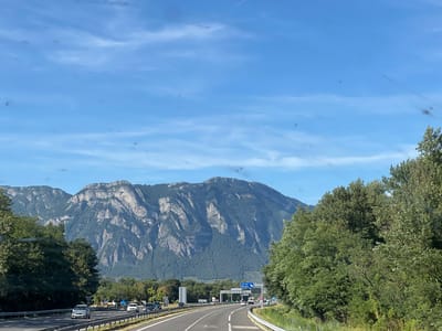 The image depicts a scenic view from a highway in Chapareillan, France, with a majestic mountain range as the backdrop. The highway, with multiple lanes, is busy with traffic, including several cars and a truck. Lush green trees line the sides of the road, leading towards the imposing mountains that dominate the horizon. The sky is a clear blue with wispy clouds, suggesting a pleasant day. The scene captures the essence of a road trip through a picturesque landscape, showcasing the beauty of the French countryside.