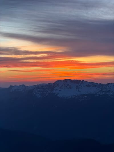 The image depicts a mountain range silhouette against a vibrant sunset sky. The location is identified as La Giettaz, France. The sky showcases vertical streaks of orange, yellow, and purple hues, typical of a sunset, while the mountain range is a dark mass with patches of white, possibly snow. The right side of the image is a dark blue or black, possibly representing the night sky or a shaded area.