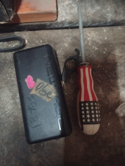 The image depicts a close-up, low-angle shot of a flat, dark-colored rectangular object, possibly a power bank, lying on a rough, textured surface. A small piece of pink material is affixed to the left side of the object, with a piece of taped paper bearing the handwritten word "OBE" in black ink. To the right of the power bank, a screwdriver with a red and white striped handle and a black end with white stars is visible. The screwdriver is positioned vertically, with its tip pointing upwards. A black pair of scissors with a curved handle is also present to the left of the screwdriver. The background consists of a textured surface that appears to be concrete or dirt, with some scattered debris. The lighting in the image is dim, casting shadows and obscuring finer details of the environment. The overall scene suggests a utilitarian setting, possibly a workshop or storage area, though no specific location within Jalingo, Nigeria can be definitively identified from the visual information provided.