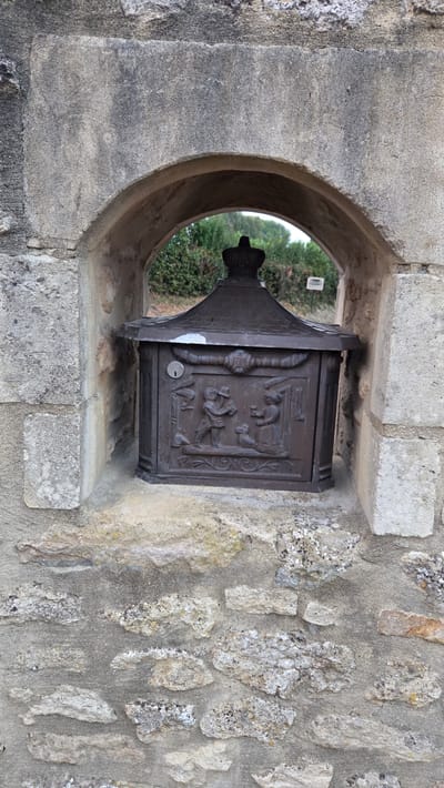 The image shows a decorative, dark metal mailbox set into an arched alcove in a stone wall. The mailbox has intricate relief carvings on its front depicting two figures, a dog, and floral motifs. A keyhole is visible above the carvings. The mailbox is topped with a peaked roof adorned with a crown-like finial. The stone wall surrounding the alcove is roughly constructed with visible mortar. In the background, through the alcove, greenery and trees are visible. A small white sign is also present in the background. The mailbox appears to be a decorative element, possibly at an entrance or on a property in Macé, France.