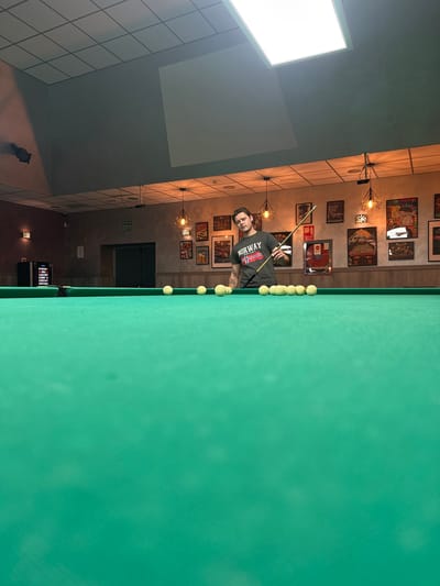 The image shows a man playing billiards in a room in San Vicente Del Raspeig/sant Vicent Del Raspeig, Spain. The billiard table is covered in green felt and has a row of white balls lined up along one edge. The man is holding a billiard cue and appears to be aiming at the balls. The room has a light-colored wall with a grid pattern and a dark-colored ceiling. There are various framed pictures and wall lamps decorating the wall. There is an emergency exit sign above the door in the background.