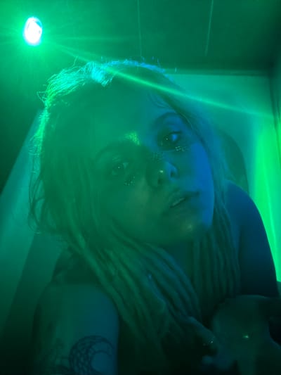 The media file is a low-angle, close-up, portrait-style photograph of a person with dreadlocks, illuminated by green and blue lights. The subject is looking directly at the camera with a neutral expression. They have glitter or some sort of metallic adornment on their forehead and cheekbones, and a chain necklace is visible. One of their hands, adorned with a ring, is near their face. A tattoo of a crescent moon with intricate patterns is visible on their arm. The background is a dimly lit, enclosed space with a bright light source in the lower left corner, casting a green glow and light flares. The overall mood is mysterious and artistic, possibly suggesting a spa, a dimly lit performance space, or a stylized photo studio. The location is identified as Vostochny District, Russia. There is no visible text in the image.