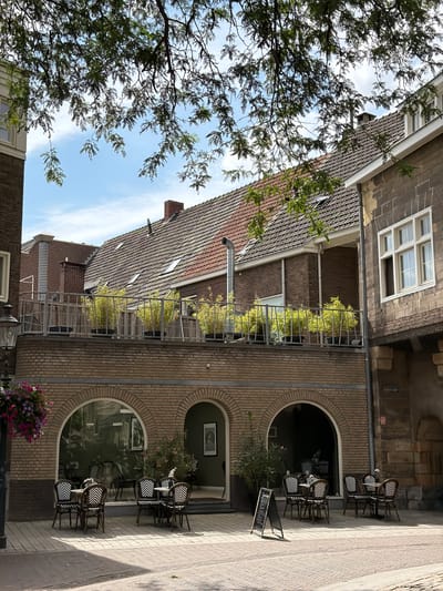 The image depicts an outdoor seating area of a cafe or restaurant in Roermond, Netherlands. The cafe is situated on a street with cobblestone paving. In the foreground, there are several tables and chairs with a black and white zigzag pattern on the upholstery. A-frame chalkboards are placed near the tables. The cafe has a brick facade with large arched windows, some of which reveal the interior. Above the cafe level, there is a balcony or terrace with metal railings, where several potted plants, likely bamboo, are displayed. In the background, there are traditional Dutch buildings with tiled roofs and dormer windows. Green foliage from a tree branches into the frame from the top, casting shadows on the scene. The sky is blue with some scattered clouds. The sign visible on the building reads "ABDHOF."