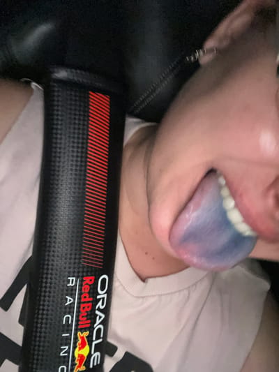 The media file shows a close-up of a person's mouth with their tongue extended. The tongue is dyed a vibrant blue and purple, with hints of pink towards the tip. The person's teeth are visible and appear white. They are wearing a light-colored t-shirt and an earring in their left ear.
In the foreground, a black object with a carbon fiber texture is visible. It features red diagonal lines on the left side and text on the right, including "ORACLE," "Red Bull," and "RACING," along with the Red Bull logo. This object appears to be a branded item, possibly related to motorsports. The overall lighting suggests an indoor setting, and no specific time of day or weather conditions can be determined. The focus is on the person's mouth and the tongue's unusual coloring, creating a visually striking and somewhat unusual composition. The presence of the Red Bull branding suggests a connection to Formula 1 racing or related events.