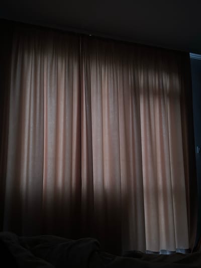 The image shows closed, horizontal blinds. The color of the blinds is a warm brown or beige. The lighting is low, creating strong shadows that accentuate the horizontal lines of the blinds. Part of a dark wall or door frame is visible on the left side of the image. The right side of the image shows a crumpled fabric, potentially a curtain or garment. The location is unknown and cannot be determined from the image. There is no information to indicate that the location is in Kribul, Bulgaria. There is no evidence of people or actions in this image.