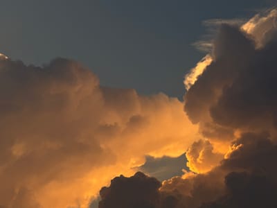 This image captures a dramatic sky filled with large, textured clouds. The lighting suggests it is either sunrise or sunset, with the sun's warm golden light illuminating the edges and undersides of the clouds, creating a striking contrast between the illuminated parts and the darker, shadowed portions. The clouds themselves appear dense and voluminous, some are a deep grey, hinting at their mass and perhaps moisture content, while others are bathed in a vibrant orange and yellow glow. A clear, deep blue sky is visible in the lower portion of the frame and peeking through gaps in the clouds. The overall impression is one of natural grandeur and power, with the interplay of light and shadow on the clouds creating a dynamic and almost sculpted appearance. There are no people or man-made objects visible, focusing solely on the atmospheric conditions. The scene evokes a sense of awe at the vastness and beauty of the sky. There is no visible text. The location context is Putten, Netherlands.