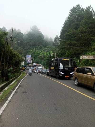 The image depicts a road scene, possibly in a mountainous or hilly area due to the presence of dense trees and fog in the background. The road has a yellow dividing line and seems congested with various vehicles, including a large black bus labeled "Mabes TNI", a gold-colored car, and several motorcycles. A billboard is visible on the left side of the road, along with utility poles and wires. The overall weather appears overcast, contributing to the foggy atmosphere. The road's surface is asphalt, and a curb with green vegetation runs alongside it. While the image is rich in contextual details, determining its precise location as Mae Chan District in Thailand isn't definitively evident from the image alone.