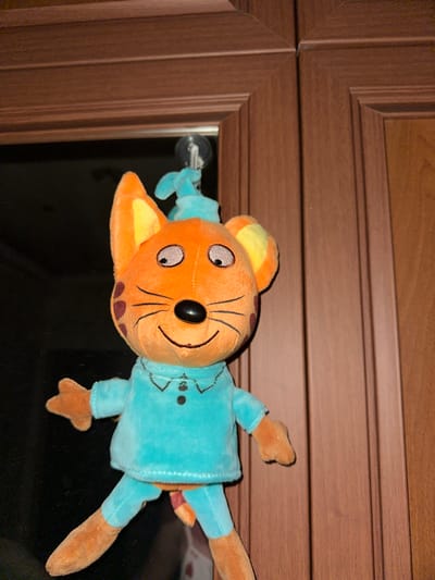 A plush toy of an orange cat character wearing a blue shirt is hanging upside down on a glass surface, possibly a door or cabinet, by a clear suction cup attached to its head. The cat has large, round eyes with black pupils and grey irises, a black button nose, and thin black lines drawn on its face to suggest whiskers and a smile. Its ears are yellow on the inside and orange on the outside, and it has brown spots on its cheeks. The toy is hanging in front of a dark, reflective surface, possibly a mirror or glass, which is mounted in a wooden cabinet. The cabinet has a dark brown, wood-paneled finish with distinct horizontal lines and beveled edges. The lighting appears to be artificial, casting a slight shadow on the plush toy. The overall scene is indoors, likely in a home or office setting. There is no visible text in the image.