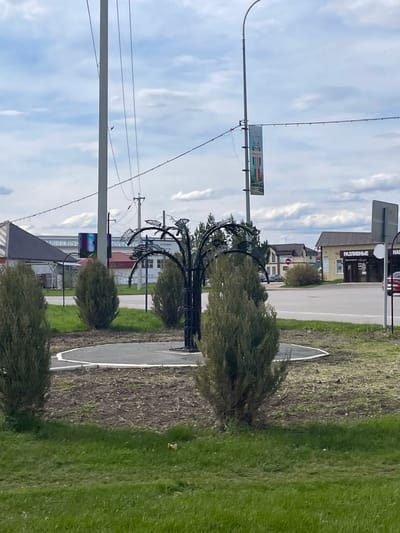 The image captures a street scene in Urussu, Russia. A sidewalk runs alongside a road, with patches of grass on either side. Small, rounded bushes line the sidewalk. A decorative, wrought-iron structure, resembling a stylized tree, stands along the sidewalk. Overhead, power lines stretch across the sky. A building with a sign in Russian is visible in the background. The sky is partly cloudy. A sign with green, white, and red colors is attached to the pole on the left.