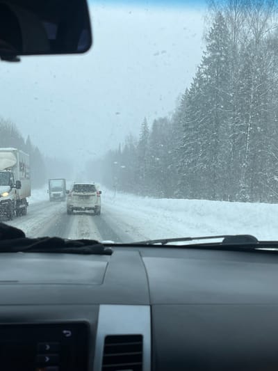 The image depicts a snowy winter road scene, viewed from the interior of a vehicle. The location is an unspecified road within or near Semenovo, Russia. Falling snow is visible through the windshield, which has water droplets or snowflakes on its surface. The dashboard of the camera vehicle is dark gray, featuring a silver trim accent on the passenger side, an infotainment screen with visible control icons on the lower left, and a central air vent. Black windshield wipers rest at the base of the windshield. A dark, soft object, possibly a glove, lies on the dashboard. A rearview mirror hangs from the ceiling, partially obscuring the upper central view.
Ahead, a snow and slush-covered two-lane road stretches into the distance. Traffic is present, with a white SUV directly ahead in the same lane, displaying accumulated snow on its rear. Behind the SUV, a smaller white utility truck with an open rear is visible. To the left, a portion of a large white truck with a box trailer is seen moving in the same direction. The roadside on both sides is banked with deep snow, and dense evergreen forests, primarily conifers and some bare deciduous trees, line the road, extending into the background under an overcast, bright gray sky. Visibility is moderately reduced due to the ongoing snowfall. No individuals are directly visible.