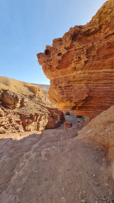 The image, taken at an unusual angle, presents a scene of rugged natural beauty in the Hevel Eilot Regional Council, Israel. The dominant feature is a massive, overhanging rock formation, characterized by its layered, reddish-brown strata, with some areas having holes or being porous, giving it a textured appearance. Below the overhanging rock is another rock formation and some soil. The angle of the photo makes it difficult to identify exactly what is going on, but you can see a road below the rock formations. A clear, blue sky forms the backdrop in the top left corner of the frame. The overall setting evokes a sense of a desert landscape.