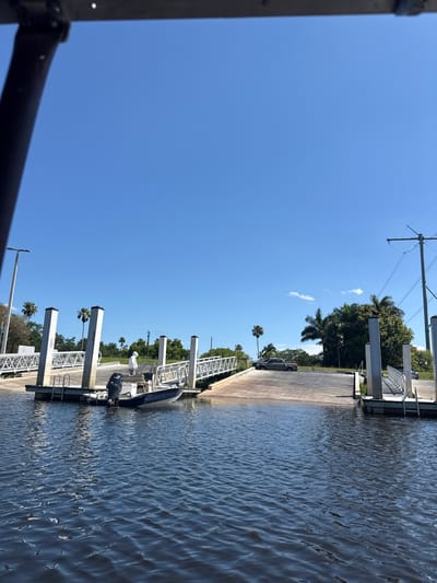 The image shows a boat ramp and dock at a waterway in Fort Lauderdale, Florida, United States. A boat is tied to the dock. The sky is clear and blue. Utility poles are visible in the background along with palm trees and other vegetation. Several cars are parked on the boat ramp.