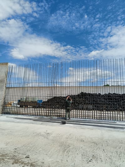 In Kladruby, Czechia, a construction site is depicted under a partly cloudy sky. The foreground shows a concrete surface with scattered debris. A large, unfinished concrete wall forms the backdrop, reinforced with a dense grid of vertical steel rebar. Behind the rebar, a large pile of black tires is visible. A person wearing a green jumpsuit is standing in front of the rebar, facing away from the camera, with their hands near their head. To the left of the tires, a blue industrial vehicle with some white components is partially visible. Along the base of the rebar structure, white tape with green lettering is affixed.