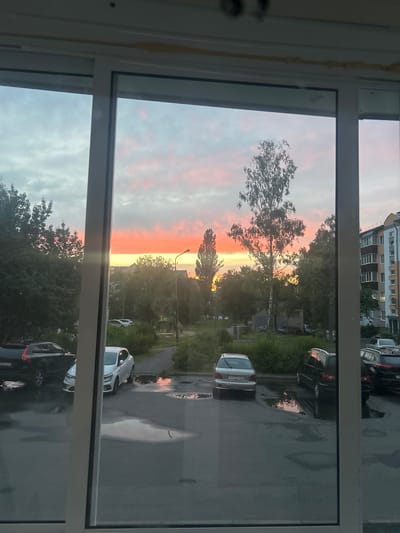 This image depicts a view through a window, likely from an apartment building in Mazyr, Belarus, overlooking an outdoor parking lot and a residential area during sunset. The sky is filled with vibrant hues of orange, pink, and yellow, indicating the sun is low on the horizon. Several trees, some tall and slender, others bushy and green, are scattered throughout the scene. A multi-story apartment building with visible windows is present in the background on the right. The parking lot is wet, with puddles reflecting the colors of the sunset and the surrounding environment. Multiple cars are parked in the lot, including a black SUV on the left, a white sedan, and other vehicles further back. There are no people visible in the frame. The foreground shows a portion of the window frame, which is white.