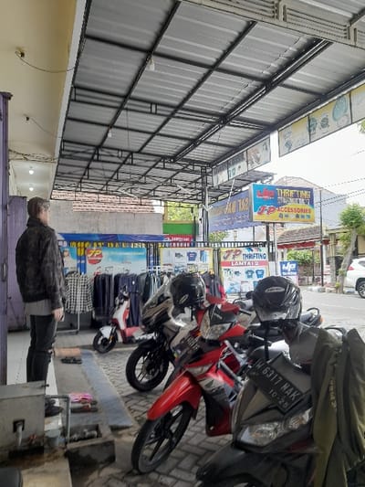 A row of parked motorbikes lines a sidewalk. The motorcycles are varying in color, with one predominantly red and others in grey. A man stands near a metal apparatus along the lower edge. In the background, several storefronts are visible, adorned with bright signs. Metal awnings extend from the building, casting shadows and creating a sense of depth in the perspective.