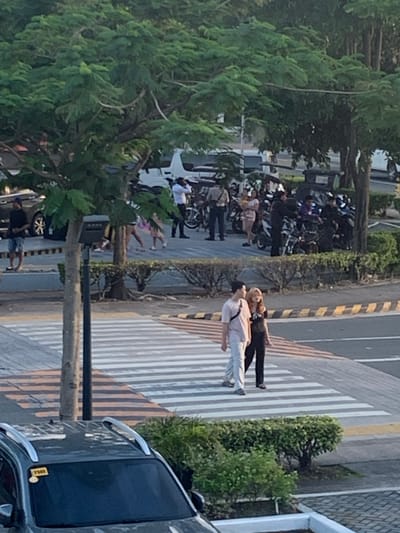 In Angeles City, Philippines, two individuals, a male and a female, are walking across a pedestrian crosswalk. The male is wearing a light-colored short-sleeved shirt and light-colored pants, while the female is wearing a dark-colored t-shirt and dark pants. They are positioned mid-stride, indicating movement across the street. In the background, several motorcycles are parked, and a group of people are visible near them. The environment appears to be an urban setting with trees and vegetation lining the street. A car is parked in the foreground.