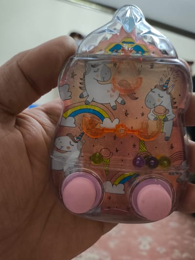 A hand holds a clear plastic toy. The toy is shaped like a plump rounded bottle and is filled with a clear liquid and colorful beads. Inside the toy, there are images of unicorns, rainbows, and ice cream cones printed on a pink background. There are also two pink, button-like objects at the bottom of the toy. The toy is held in front of a plain, light-colored wall. There is no visible connection to the city of Khasavyurt, Russia.