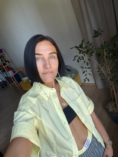 In Postol, Russia, a woman with dark bobbed hair and blue eyes takes a selfie in what appears to be a domestic indoor setting. She is wearing a light yellow open button-up shirt over a black bralette, revealing her midriff. Her pants are gray with thin white vertical stripes, and a label on the waistband reads "BE YOURSELF IN my @plains CLOTHES." A silver necklace with a pendant hangs around her neck. She is also wearing a gold wristwatch on her left wrist. The background includes a tall potted plant with green leaves, a light-colored curtain, and shelves with various items. The lighting suggests it is daytime, with light streaming in from the left. The floor is wooden.
