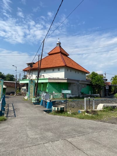 The scene is a street corner in Kecamatan Tulangan, Indonesia, located at latitude: -7.475, longitude: 112.616. A small mosque, with an orange tiled roof and white walls, is visible on the right side of the street. The mosque is located behind a low stone wall, with a green painted fence. On the left side of the street is a concrete bridge, with a metal gate. There is a sign indicating the street name, "JL. LAKSANA." There is a small metal pole on the left side of the bridge, with a sign, "RW. 02," visible on the side. The street is paved with concrete and there are power lines running overhead. The sky is blue with white clouds, and there are trees and shrubs visible in the background. There is a small, shallow stream on the other side of the wall from the mosque.  There are no people present.