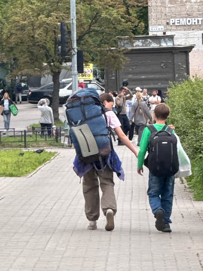 In Канонерец Снт, Russia, two individuals are walking on a paved sidewalk. The person on the left, with a large blue backpack, is wearing dark pants and white shoes. They are walking away from the camera. The person on the right, a younger individual, is wearing blue jeans and dark shoes, and also carries a black backpack and a white bag. They are walking in the same direction. Behind them, a group of people are visible in the background, some of whom appear to be looking at something or interacting. Trees and foliage are present along the side of the sidewalk, and a building with brickwork is visible in the distance. A traffic light is mounted on a pole to the left of the frame.