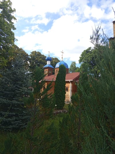 A wooden church with blue domes and golden crosses stands amidst lush greenery. The church has a red roof and visible windows. Tall evergreen trees and deciduous trees surround the structure. The sky is partly cloudy with blue patches. The scene appears to be located in a serene, possibly rural or park-like setting within Giaginskaya, Russia. There are no people visible in the image.